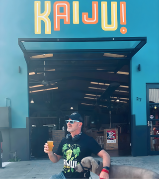 Man sitting on a bench with a dog, holding a drink, in front of a building with 'KAIJU!' branding.